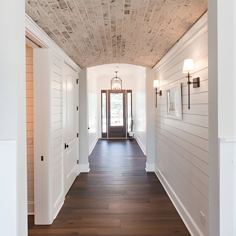 Elegant hallway with brick ceiling, dark wood floor, and white shiplap walls, leading to a glass front door. Designed by a home builder.