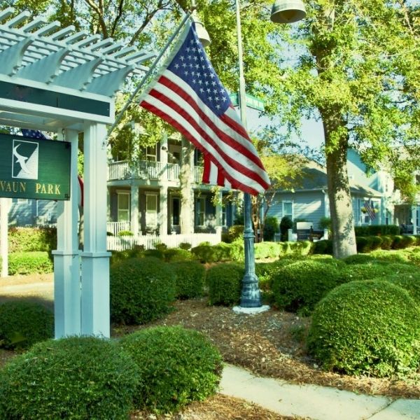 Peaceful park scene with American flag, manicured shrubs, and charming homes in background, highlighting community focus by home builders.