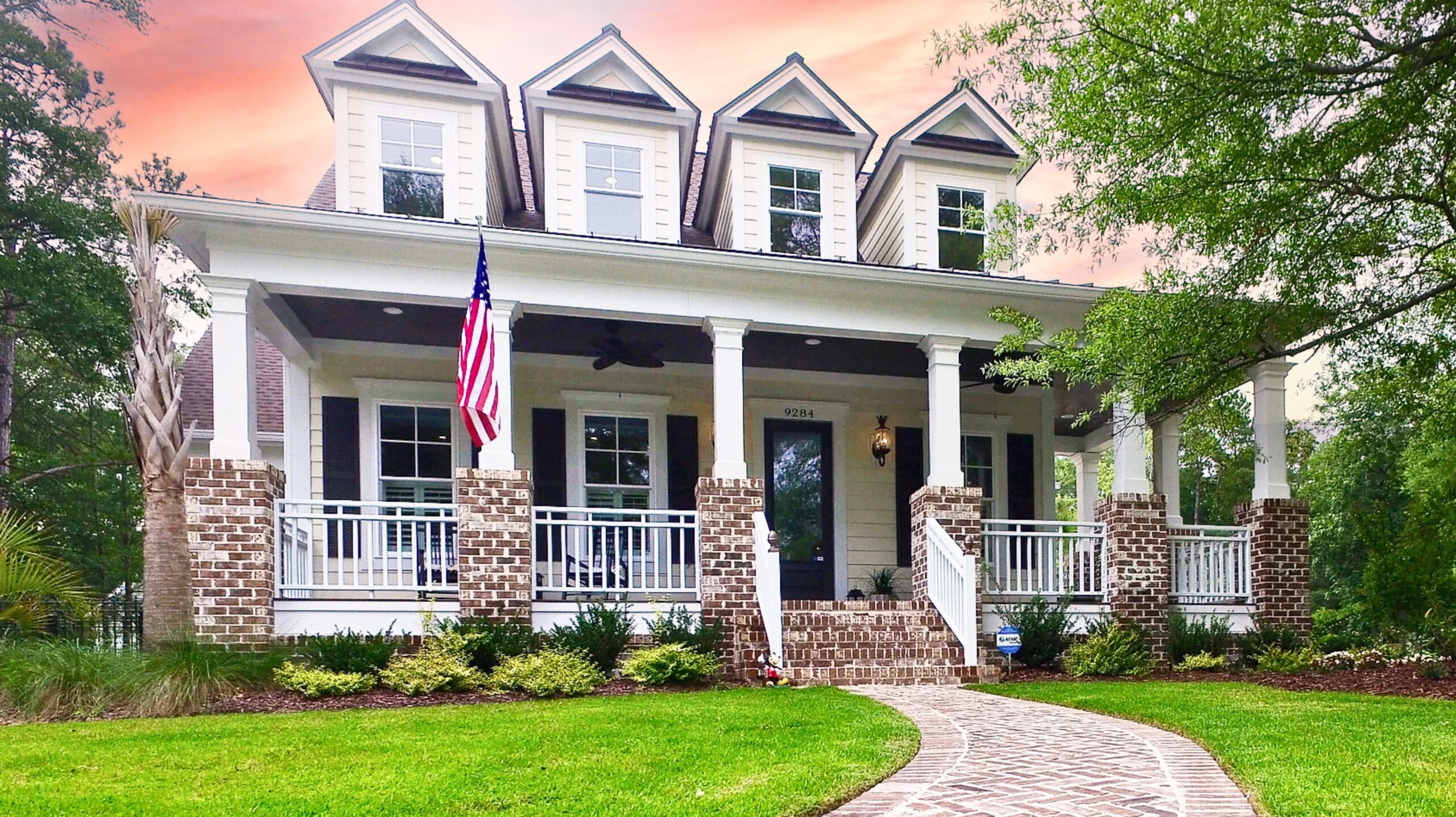 Charming colonial-style home with wraparound porch and American flag, showcasing the craftsmanship of a home builder. Beautiful sunset backdrop.