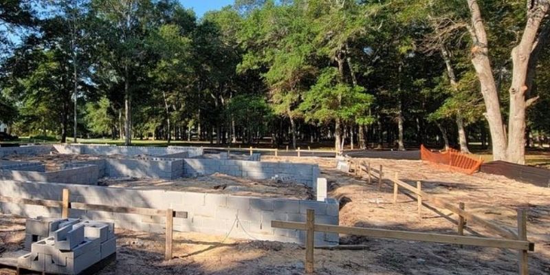 Foundation of new home under construction by home builder, surrounded by wooded area and open sky.