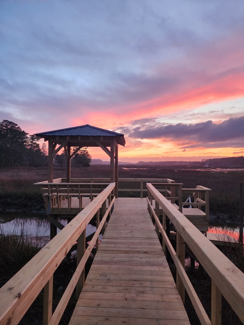 Sunset over a wooden dock with railing and gazebo, built by a home builder, stretching over serene marshland.