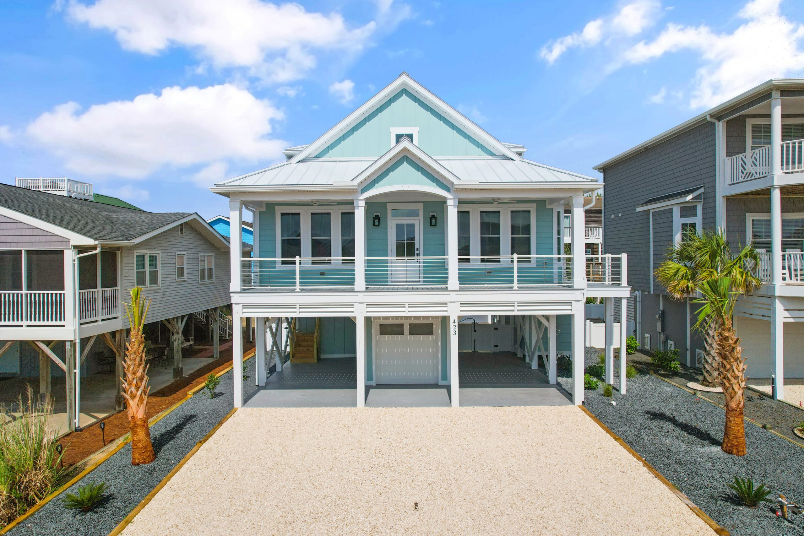 Elevated light blue house with white accents, featuring a spacious porch and palm trees, exemplifying modern coastal home builder design.