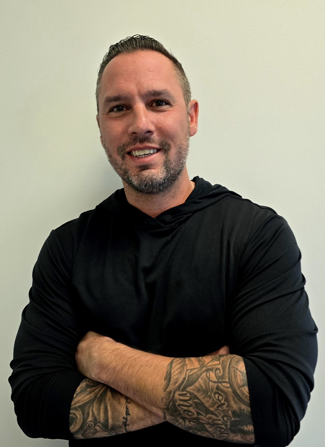 Smiling man in black shirt with tattooed arm crossed, standing against a plain backdrop.