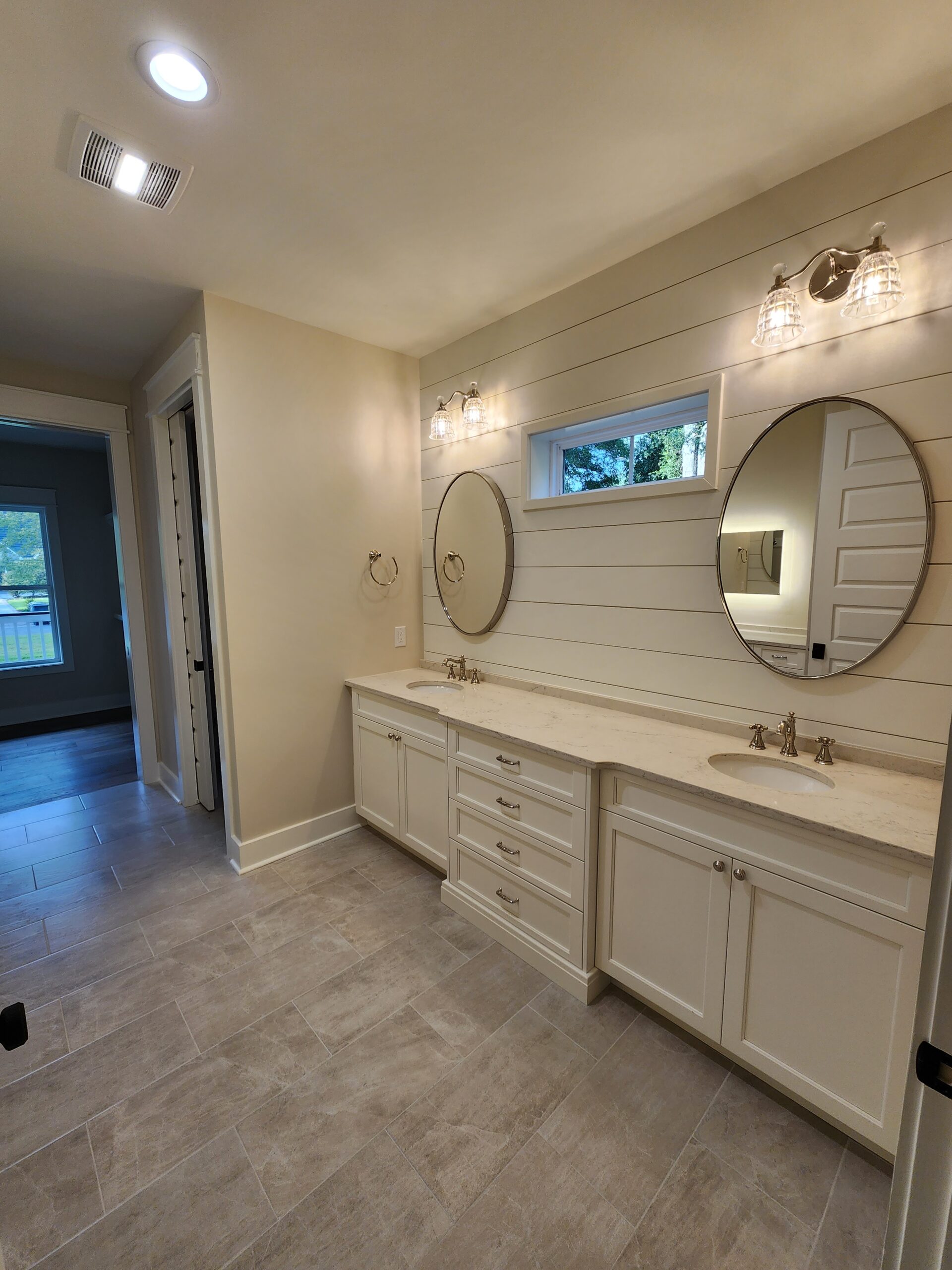 OceanHarbourEstates (4) Modern bathroom by home builder featuring dual sinks, round mirrors, beige tile floor, and sleek lighting above a shiplap wall.