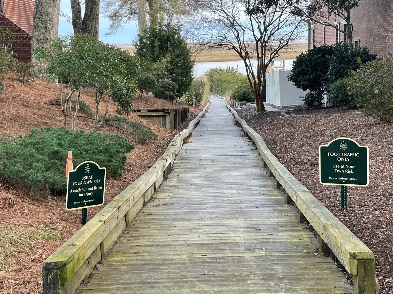 Wooden boardwalk flanked by greenery and caution signs, leading to a water view. Ideal path design for a home builder project.